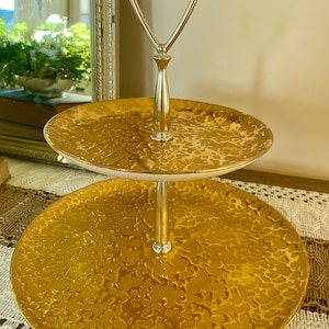 May include: A two-tiered serving tray with a gold textured finish. The tray has a silver handle and a central support column. The plates have a hammered texture. The tray is designed for serving desserts or appetizers.