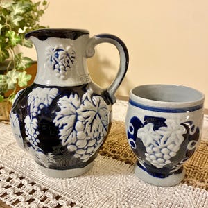 May include: A blue and white ceramic pitcher and matching cup. The pitcher has a handle and is decorated with grape and leaf designs. The cup is short and round, also with grape and leaf designs. The pitcher has the number "0.25" on it.
