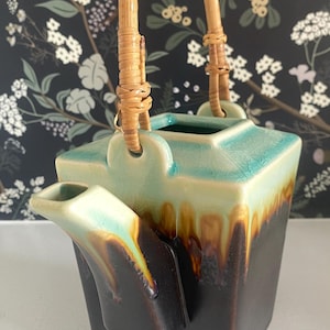 May include: A square ceramic teapot featuring a dark brown base with a light turquoise glaze dripping down from the top rim. The teapot has a curved spout and a tall, arched handle made of woven light brown rattan attached to two side loops.
