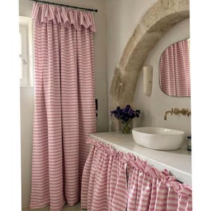 Puede incluir: Una cortina de cuerpo entero y un faldón de lavabo a juego con un patrón de rayas horizontales en rosa y blanco. Ambas piezas presentan un detalle de volantes fruncidos en el borde superior, creando un aspecto coordinado.