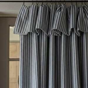 May include: A window curtain featuring a vertical striped pattern in dark blue and off-white. The fabric hangs from a black rod using metal rings, creating deep, gathered folds along the top header that extend down the length of the panel.