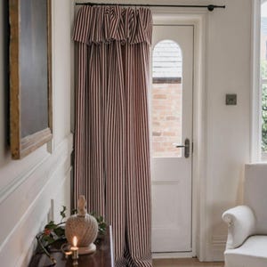 May include: A floor-length curtain featuring a vertical striped pattern in dark red and white, suspended from a black metal rod. The curtain includes a ruffled valance at the top and hangs over a white door with an arched glass window pane.