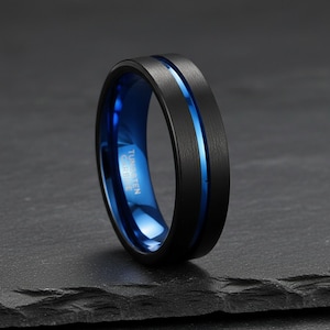 Könnte beinhalten: Ein mattschwarzer Ring aus Wolframkarbid mit gebürsteter Oberfläche außen und polierter blauer Innenseite. Eine dünne, vertiefte blaue Rille verläuft horizontal um die Mitte des Rings. Der Text TUNGSTEN CARBIDE ist auf der Innenseite eingraviert.