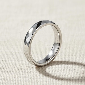Può includere: Un anello in carburo di tungsteno argentato con un design sfaccettato. L'anello ha una finitura lucida e il testo "TUNGSTEN CARBIDE" è inciso all'interno. L'anello è mostrato su uno sfondo neutro.