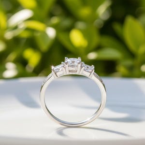 May include: A silver ring with a three-stone design. The center stone is round, flanked by two smaller, rectangular stones. The band is also adorned with smaller stones. The ring is set against a blurred green background.
