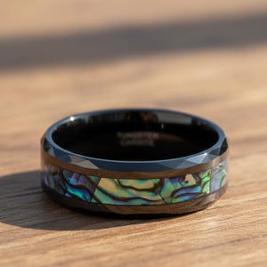 May include: Black tungsten carbide ring with a faceted outer edge and a band of iridescent abalone shell. The ring is set on a wooden surface, showcasing its polished finish and unique design.
