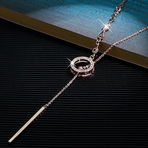 Puede incluir: Collar de oro rosa con un colgante circular adornado con cristales transparentes. El collar presenta una delicada cadena y un colgante de barra lineal. La joya se exhibe sobre un fondo oscuro.