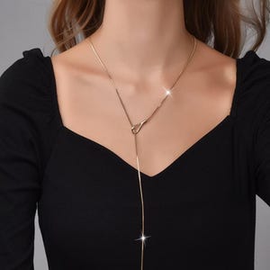 Può includere: Una collana color oro con una catena delicata e un design a forma di Y. La collana presenta un piccolo anello nella parte superiore e una catena più lunga e pendente che termina a goccia. La collana è indossata su un top nero.