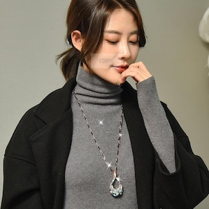 Op de afbeelding: Een vrouw draagt een grijze coltrui en een zwarte jas. Een lange ketting met een druppelvormige hanger met blauwe en heldere kristallen is zichtbaar. De afbeelding heeft de tekst "100%" in het midden.