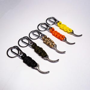 Puede incluir: Cinco llaveros con cordón trenzado en negro, verde oliva, beige, naranja y amarillo. Cada llavero tiene un mosquetón plateado y un gancho de metal curvado. Los llaveros están dispuestos en diagonal sobre un fondo blanco.