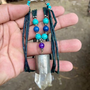 May include: A handmade necklace featuring a clear quartz crystal pendant with a silver-colored metal cap. The necklace has turquoise, blue, and purple beads, and dark blue string. The necklace is held in a hand.