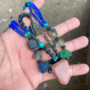 May include: A handmade necklace featuring a variety of colorful beads and stones. The necklace includes blue, turquoise, and green beads, along with larger, irregularly shaped stones in shades of pink, blue, and gray. The necklace is held in a hand.