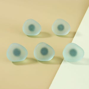 Op de afbeelding: Vijf lichtblauwe, kiezelvormige decoratieve objecten met een donkerder blauw centrum. De objecten zijn gerangschikt op een lichtgele en witte ondergrond. De objecten hebben een matte uitstraling.