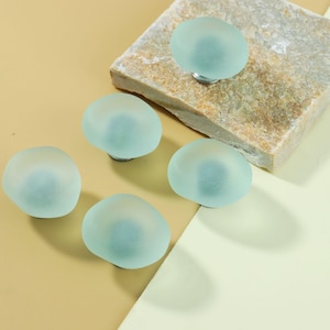 May include: Five frosted seafoam green cabinet knobs with silver hardware. The knobs have a smooth, rounded shape, resembling small stones. One knob is attached to a textured, rectangular stone. The knobs are arranged on a light tan and cream colored surface.