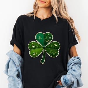 Puede incluir: Camiseta negra con un gran diseño de trébol. El trébol consta de tres hojas verdes en forma de corazón con detalles florales blancos, una herradura dorada y detalles de nudos celtas. El diseño está cosido con hilo dorado.