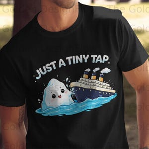 May include: Black t-shirt featuring a cartoon iceberg with a smiling face, colliding with a cruise ship. The text "JUST A TINY TAP" is above the illustration. The design is in blue, white, and yellow.