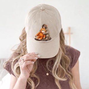 以下が含まれることがあります： ベージュ色の野球帽。2匹の猿が抱き合っている刺繍デザインが施されています。1匹はオレンジ色、もう1匹はグレー色です。帽子にはカーブしたつばと、上部にボタンが付いています。帽子はニュートラルカラーです。