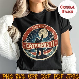 Puede incluir: Camiseta negra con un diseño circular desgastado que representa a un astronauta alcanzando la luna, con el texto "MISSION CATERMIS II". El diseño utiliza elementos rojos, azules y blancos, con estrellas y detalles de planetas.