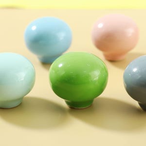 May include: A collection of colorful ceramic knobs in various pastel shades, including blue, green, and pink. Each knob has a rounded top and a short, cylindrical base. The knobs are arranged on a light yellow surface.