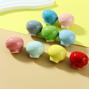 Może przedstawiać: Kolekcja dziewięciu kolorowych ceramicznych gałek w różnych odcieniach, w tym niebieskim, różowym, zielonym, żółtym i czerwonym. Gałki mają zaokrągloną górę i krótką, cylindryczną podstawę. Ułożone są na żółtej i beżowej powierzchni.