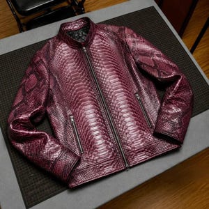May include: A deep purple jacket featuring a textured snakeskin pattern throughout. The garment includes a central full-length zipper, a stand-up collar, and two zippered side pockets. The interior lining is black, and the cuffs are finished with a straight hem.