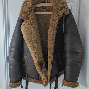 Può includere: Giacca bomber in pelle marrone con colletto e fodera in montone. La giacca ha una chiusura con cerniera, dettagli con fibbie e un aspetto vintage e invecchiato. La giacca è appesa a una gruccia di legno.