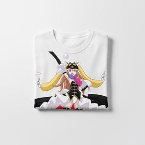 May include: White t-shirt featuring a colorful anime-style illustration. The design includes a character with long blonde hair, a black and white outfit, and a pink heart-shaped detail. The shirt is folded neatly.