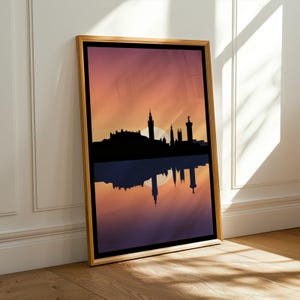 Peut inclure: Une impression d'art mural verticale encadrée présentant la silhouette noire d'une ligne d'horizon urbaine sur un ciel au coucher du soleil dégradé orange et violet. L'image inclut un reflet inversé de la ville dans la moitié inférieure.