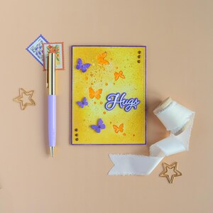 May include: A greeting card with a yellow background and purple border, decorated with orange and purple butterflies. The word "Hugs" is written in cursive. A purple pen, stamps, ribbon, and star-shaped paper clips are also present.