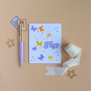 May include: A white card with butterfly cutouts and the word "Hugs" in purple script. The card is decorated with orange, yellow, and purple butterflies. A purple pen, stamps, ribbon, and star-shaped paper clips are also present.