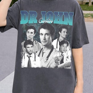 May include: Dark gray t-shirt featuring a vintage design with the text "DR JOHN Carter" in a teal and blue gradient. The shirt displays multiple black and white images of a man wearing a stethoscope and medical attire.