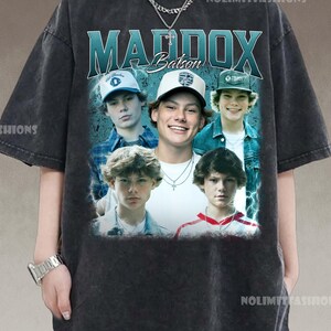 May include: A dark gray t-shirt featuring a graphic of the name "MADDOX Batson" in teal and white, with a collage of five young men wearing baseball caps. The shirt has a vintage, distressed look.