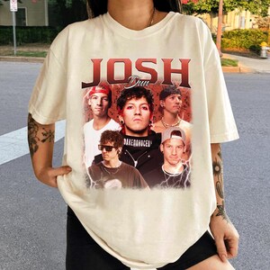 May include: Cream-colored t-shirt featuring a graphic of the band "Josh Dun" with a vintage design. The design includes images of the band members. The text "Josh Dun" is in a bold, red font.