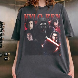 May include: Dark gray t-shirt featuring a graphic of Kylo Ren from Star Wars. The design includes multiple images of the character, with the name "KYLO REN" in red block letters at the top. The shirt is a casual, oversized style.