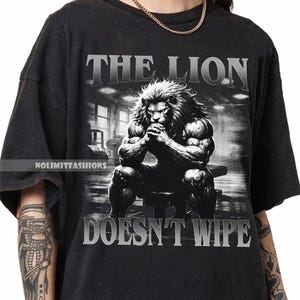 May include: Black t-shirt featuring a muscular lion illustration in a gym setting. The text "THE LION DOESN'T WIPE" is printed above and below the lion image. The shirt has a crew neck and short sleeves.