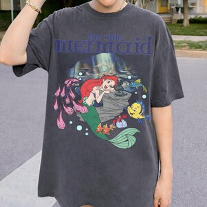 Pu&ograve; includere: T-shirt grigio scuro con una grafica di Ariel de La Sirenetta. Il design include Ariel con i capelli rossi e una coda verde, circondata da pesci e la scritta "the little MERMAID" in blu.
