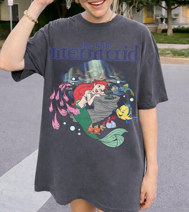 Pu&ograve; includere: T-shirt grigio scuro con una grafica di Ariel de La Sirenetta. Il design include Ariel con i capelli rossi e una coda verde, circondata da pesci e la scritta "the little MERMAID" in blu.