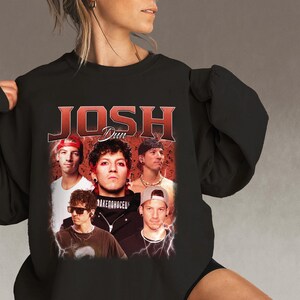 May include: Black crew neck sweatshirt featuring a vintage-style graphic of Josh Dun with the text "JOSH DUN" in red. The graphic includes multiple images of the artist. The sweatshirt is a dark color.