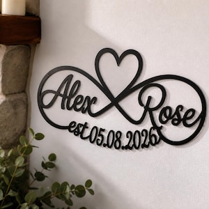 May include: A black metal wall sign featuring an infinity symbol intertwined with a heart shape. The sign displays the names Alex and Rose in a cursive script, with the text est 05.08.2026 positioned below the names within the infinity loop.