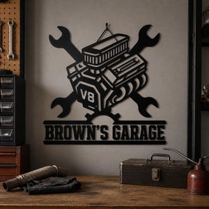 May include: A black metal wall sign featuring a V8 engine graphic centered over two crossed wrenches. Below the engine, a gear icon sits above a rectangular banner with the text BROWN'S GARAGE in bold, capitalized, sans-serif lettering.