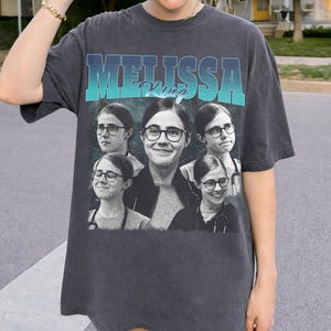 May include: Dark grey oversized t-shirt with the name "MELISSA King" in a teal and blue gradient font. The shirt features a collage of images of a person wearing glasses and a stethoscope.