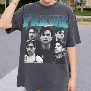 Op de afbeelding: Donkergrijs t-shirt met een afbeelding van een man en de naam "FRANK Langdon" in blauw en turquoise. De afbeelding bevat meerdere afbeeldingen van dezelfde man. Het shirt heeft een vintage stijl.
