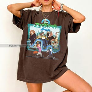 May include: A brown t-shirt with a graphic from the Disney film "Zootopia 2." The design includes the film's title and several animal characters. The shirt has short sleeves and a loose fit.