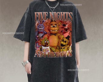 May include: Dark gray t-shirt featuring a graphic from the video game "Five Nights at Freddy's." The design includes several animated characters and the game's title in a distressed, vintage style. The shirt has short sleeves and a crew neck.