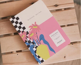 May include: A journal with a pink and cream cover featuring floral and geometric designs. The journal's cover has the text "BLESSED BETTER" and "THE QURAN JOURNAL" with the brand name "HALISAN". The journal is resting on a wooden surface.
