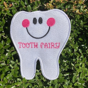 May include: White felt tooth with a smiling face, pink cheeks, and black eyes. The text "TOOTH FAIRY" is embroidered in pink on the tooth.