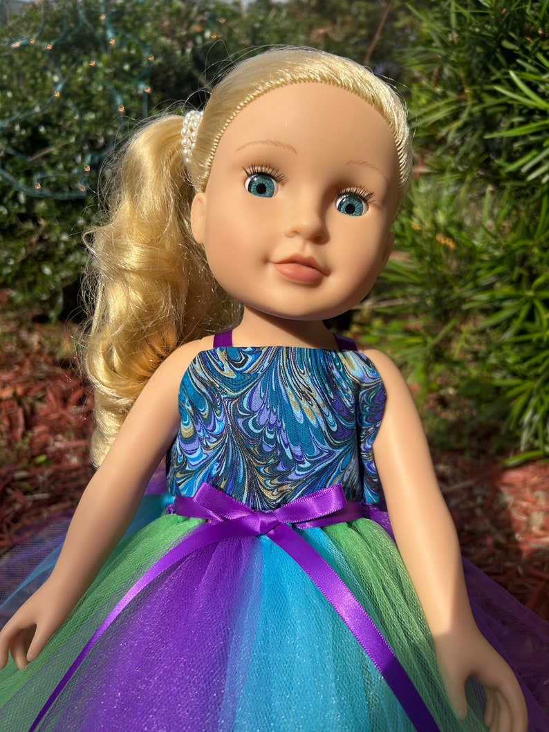 May include: A doll wearing a blue and green marbled print dress with a purple tulle skirt and a purple satin bow.
