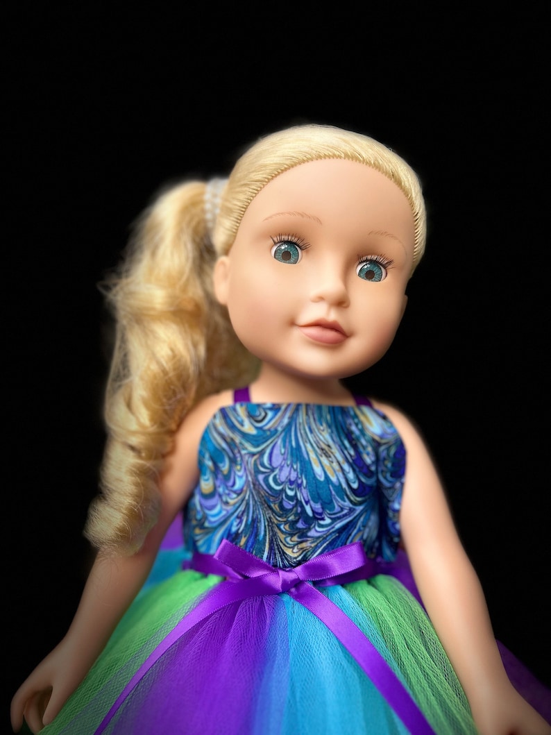 May include: A doll with blonde hair wearing a blue and gold patterned dress with a purple tulle skirt and a purple satin bow.