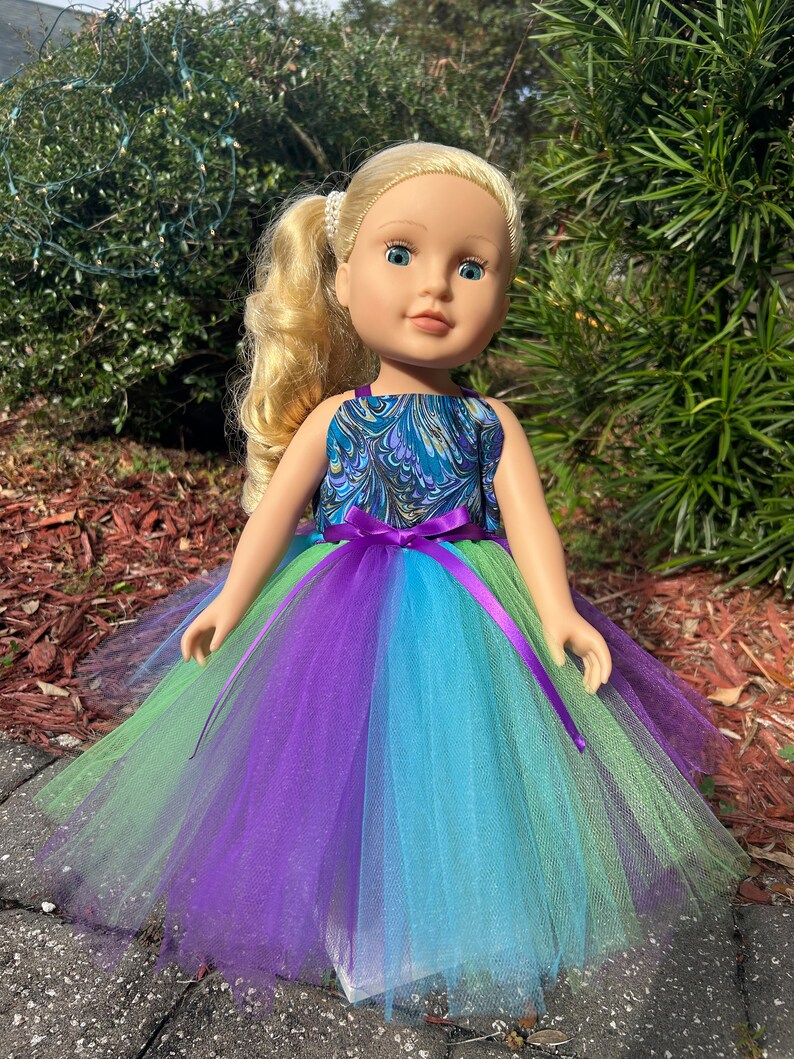 May include: A doll wearing a teal, purple, and green tulle dress with a purple ribbon. The doll has blonde hair and blue eyes.