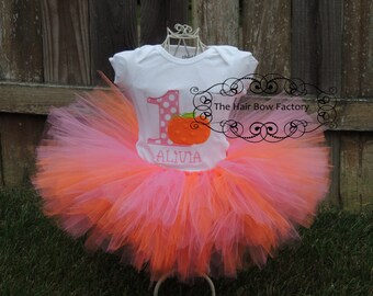 May include: A white onesie with a pink number 1 and a pumpkin graphic, paired with a layered pink and orange tutu. The onesie has the name "ALIVIA" embroidered below the pumpkin. The Hair Bow Factory logo is visible.
